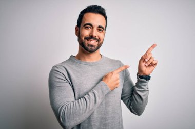 Sakallı, sıradan bir süveter giyen, beyaz arka planda duran, gülümseyen ve kameraya bakan iki eli ve parmakları yandan gösteren genç yakışıklı bir adam..