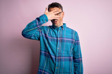 Gündelik gömlek ve gözlük takan genç yakışıklı adam izole edilmiş pembe arka planda dikiliyor. Şok olmuş bir şekilde yüzünü ve gözlerini kapıyor ve utanmış bir ifadeyle parmaklarından bakıyor..