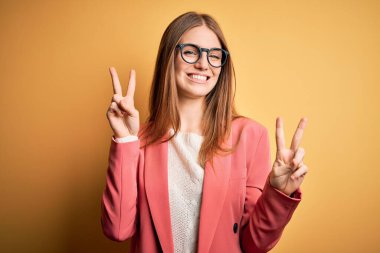 İzole edilmiş sarı arka planda gözlüklü, ceketli, güzel kızıl saçlı kadın kameraya bakıp zafer işareti yaparken gülümsüyor. İki numara..