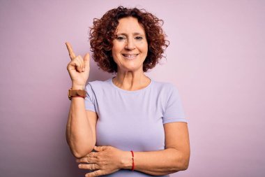Orta yaşlı güzel kıvırcık saçlı kadın izole edilmiş pembe arka planda günlük tişört giyiyor yüzünde kocaman bir gülümseme, eli ve parmağı kameraya bakıyor..