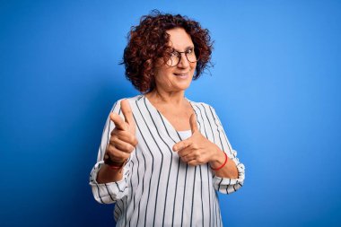 Orta yaşlı, kıvırcık saçlı, sıradan çizgili tişörtlü, izole edilmiş arka plan üzerinde mutlu ve komik yüzlü parmakları kameraya gösteren güzel bir kadın. İyi enerji ve titreşimler.