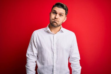 Mavi gözlü, zarif gömlekli genç bir iş adamı. Kırmızı izole edilmiş arka planda duruyor. Komik yüzüyle yanaklarını şişiriyor. Havayla şişmiş ağız, çılgın bir ifade..