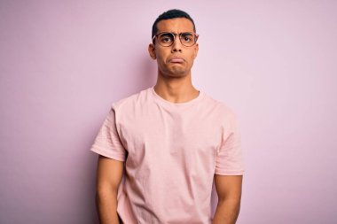 Yakışıklı Afro-Amerikan erkeği pembe arka planda günlük tişört ve gözlük takıyor. Depresyonda ve endişe içinde, sinirli ve korkmuş. Üzücü ifade.