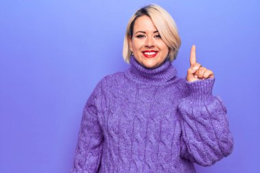Güzel sarışın ve cüsseli bir kadın mor arka planda balıkçı yaka kazak giyiyor. Bir numarayı işaret ediyor ve kendinden emin ve mutlu gülümsüyor..
