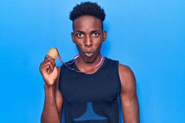 Afro-Amerikalı genç adam spor giysisi takıyor. Elinde madalya var. Korkmuş ve ağzı açık bir şekilde hayrete düşmüş. 
