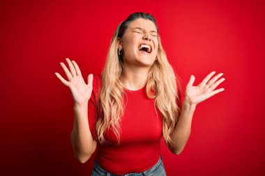 İzole edilmiş kırmızı arka plan üzerinde dikilmiş, kollarını kaldırmış ve gözleri kapalı heyecanlı bir şekilde çılgın ve çılgın bir şekilde başarıyı kutlayan genç, sarışın bir kadın. Kazanan konsept