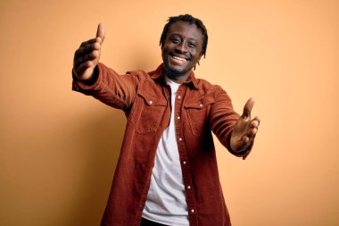 Sarı arka planda sıradan bir ceket giyen genç ve yakışıklı Afro-Amerikan adam sarılmak için kollarını açmış gülümseyen kameraya bakıyor. Mutluluğu kucaklayan neşeli bir ifade..