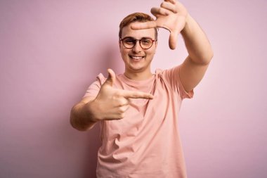 Genç, kızıl saçlı, günlük tişört giyen, pembe arka planda duran, gülümseyen, elleri ve parmakları mutlu yüzlü genç bir adam. Yaratıcılık ve fotoğrafçılık kavramı.