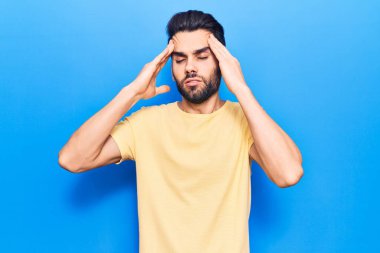 Sakallı, elleri kafasında, günlük tişört giyen genç yakışıklı bir adam. Stres yüzünden baş ağrısı çekiyor. migren ağrısı. 