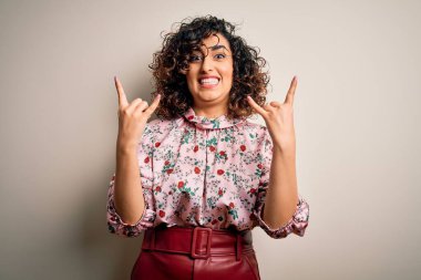 Güzel kıvırcık Arap kadın çiçek tişörtü giyip izole edilmiş beyaz arka planda duruyor çılgın bir ifadeyle elleri havada rock sembolü yapıyor. Müzik yıldızı. Ağır konsept.