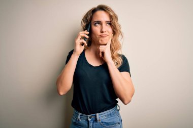 Güzel sarışın kadın beyaz arka planda akıllı telefondan konuşuyor. Ciddi bir yüz ifadesi var. Soru sormayı düşünüyor.