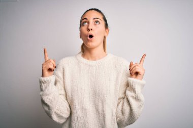 Mavi gözlü genç sarışın kadın beyaz arka planda sıradan bir süveter giyiyor şaşkın şaşkın yukarı bakıyor ve parmaklarıyla işaret ediyor ve kollarını kaldırıyor..