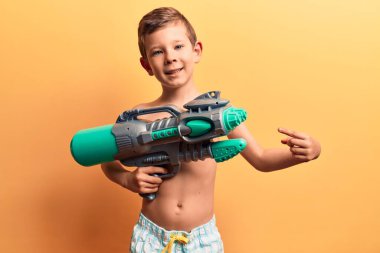 Mayo giyen tatlı sarışın çocuk elinde su tabancası, gülümsüyor mutlu eli ve parmağı ile işaret ediyor. 