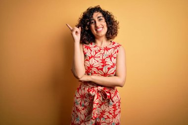 Genç, kıvırcık Arap bir kadın tatilde. Çiçekli bir elbise ve yüzünde kocaman bir gülümseme olan güneş gözlüğü takıyor. Elleri ve parmakları kameraya bakıyor..