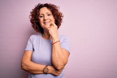 Orta yaşlı güzel kıvırcık saçlı kadın pembe arka plan üzerine günlük tişört giyip kameraya bakarken kollarını kavuşturup gülümsüyor ve çenesini kaldırıyordu. Olumlu düşünüyorum..