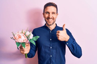 Genç, yakışıklı, romantik bir adam pembe arka planda çiçeklerle dolu bir buket tutuyor mutlu ve pozitif gülümsüyor, başparmağıyla mükemmel ve onay işareti yapıyor.