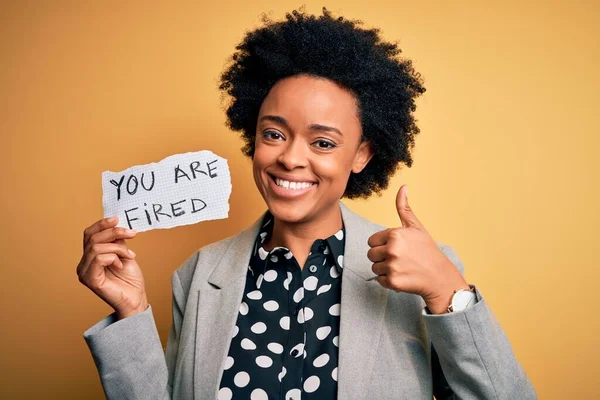 Young African American afro woman with curly hair holding paper with you are fired message happy with big smile doing ok sign, thumb up with fingers, excellent sign