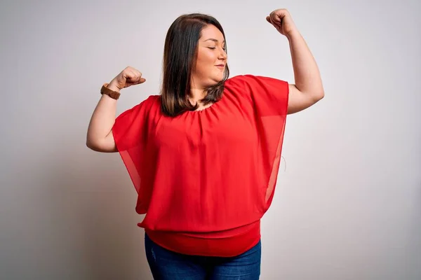 Beautiful brunette plus size woman wearing casual red t-shirt over isolated white background showing arms muscles smiling proud. Fitness concept.