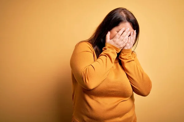 Beautiful brunette plus size woman wearing casual sweater over isolated yellow background with sad expression covering face with hands while crying. Depression concept.