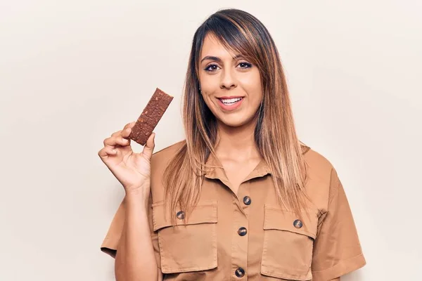 Young beautiful woman eating energy protein bar looking positive and happy standing and smiling with a confident smile showing teeth