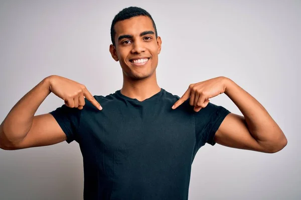 若いハンサムなアフリカ系アメリカ人男性は 笑顔で自信を持って白い背景に立ってカジュアルなTシャツを着て 誇りに思って幸せな指で自分自身を指して — ストック写真