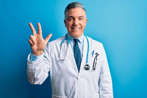 中年英俊 白发苍苍的医生 身穿外套 头戴蓝色听诊器 三指一指 面带微笑 自信而快乐 — 图库照片