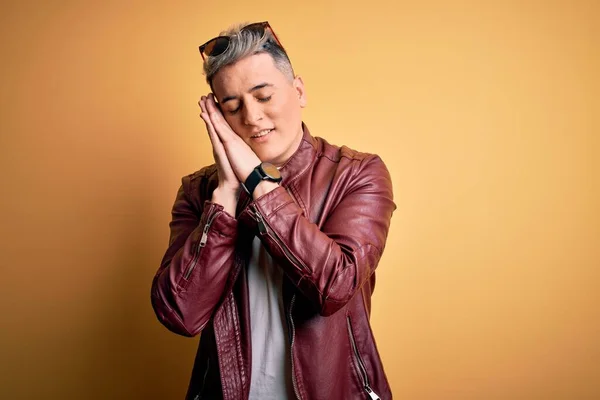 Young handsome modern man wearing fashion leather jacket and sunglasses over yellow background sleeping tired dreaming and posing with hands together while smiling with closed eyes.