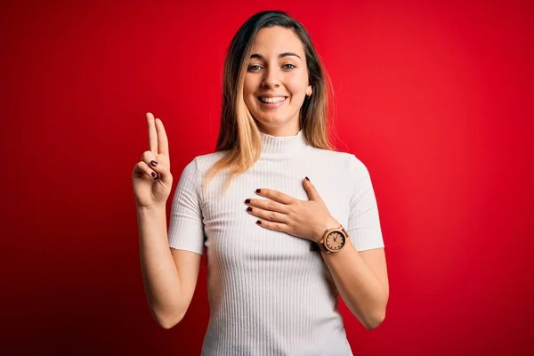 Beautiful blonde woman with blue eyes wearing casual white t-shirt over red background smiling swearing with hand on chest and fingers up, making a loyalty promise oath