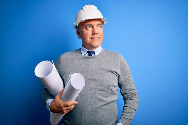 Middle age handsome grey-haired architect man wearing safety helmet holding blueprints smiling looking to the side and staring away thinking.