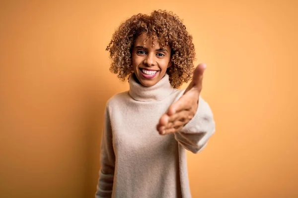 黄色の背景にタートルネックセーターを身に着けている若い美しいアフリカ系アメリカ人女性は 挨拶や歓迎として握手を提供しています 成功事業 — ストック写真