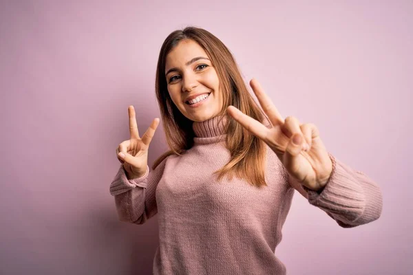 Beautiful young woman wearing turtleneck sweater over pink isolated background smiling looking to the camera showing fingers doing victory sign. Number two.