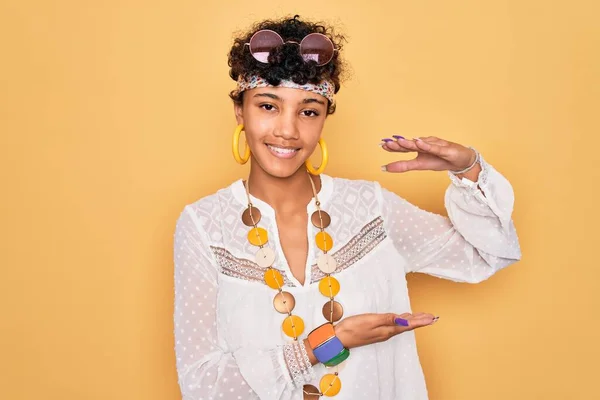Young beautiful african american afro hippie woman wearing sunglasses and accessories gesturing with hands showing big and large size sign, measure symbol. Smiling looking at the camera. Measuring concept.