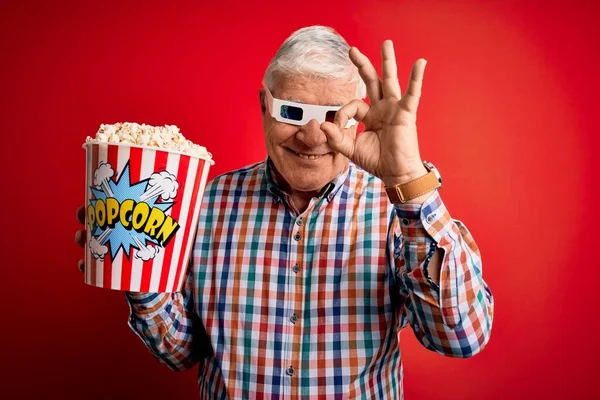 Senior hoary man watching film using 3d glasses eating popcorn over red background doing ok sign with fingers, excellent symbol