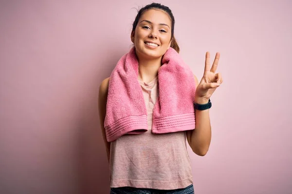 若い美しいブルネットのスポーツウェアとタオルを身に着けている女性ピンクの背景に表示され 指の数2で指摘しながら 自信と幸せ — ストック写真