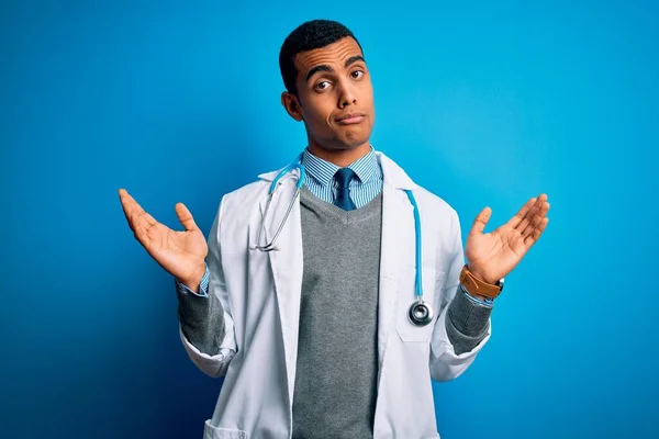 青い背景の上にコートと聴診器を身に着けているハンサムなアフリカ系アメリカ人医師の男性は 腕や手で上げられ 混乱した表情 疑いの概念 — ストック写真
