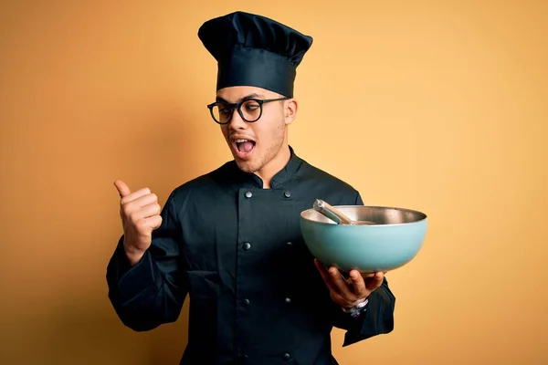 Young brazilian chef man wearing cooker uniform and hat using bowl and whisk pointing and showing with thumb up to the side with happy face smiling