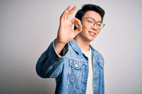 Young handsome chinese man wearing denim jacket and glasses over white background smiling positive doing ok sign with hand and fingers. Successful expression.