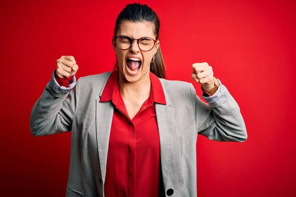 赤を背景にジャケットやメガネを身に着けている若い美しいブルネットの実業家は怒りで叫んでいる間に怒りと怒りの拳を上げ怒っている 激怒と積極的な概念 — ストック写真