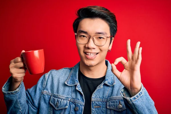 Young handsome chinese man drinking cup of coffee over isolated red background doing ok sign with fingers, excellent symbol