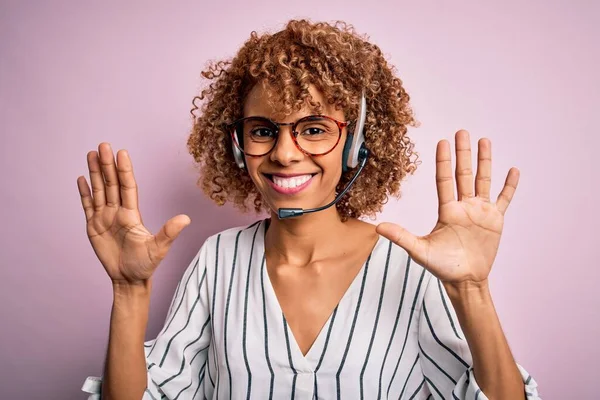 African american curly call center agent woman working using headset over pink background showing and pointing up with fingers number ten while smiling confident and happy.
