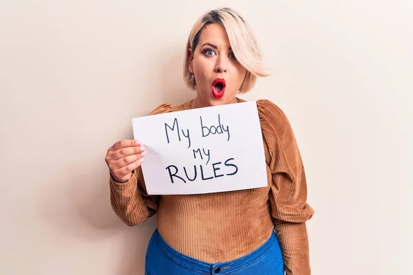 Blonde plus size woman asking for women rights holding paper with my body my rules message scared and amazed with open mouth for surprise, disbelief face