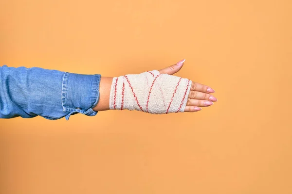 Wounded hand of caucasian young woman cover with bandage doing handshake gesture over isolated yellow background