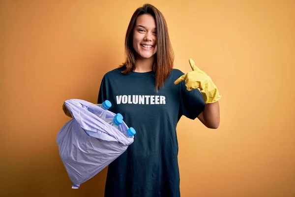 Beautiful volunteer girl caring environment doing volunteering holding bag with rubish bottles with surprise face pointing finger to himself