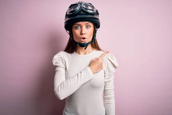 若いです美しいですオートバイの女性とともに青い目ピンクの背景の上に元ヘルメットを身に着けています側面に指で指差し込み 口を開けて驚きの表情 — ストック写真