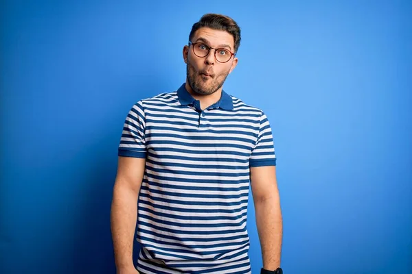 Young man with blue eyes wearing glasses and casual striped t-shirt over blue background making fish face with lips, crazy and comical gesture. Funny expression.