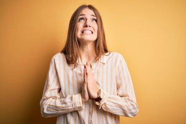 İzole edilmiş sarı arka plan üzerinde sıradan çizgili tişört giyen genç, güzel, kızıl saçlı bir kadın elleri bağlı yalvarıyor ve dua ediyor. Yüzünde umut ifadesi var. Çok duygusal ve endişeli. Yalvarıyorum..