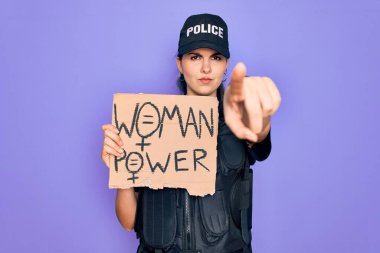 Güvenlik kurşungeçirmez yelek giyen kadın polis memuru elinde kadın güç gösterisi kartonuyla kamerayı işaret ediyor ve sen, el işareti, ön taraftan pozitif ve kendinden emin bir hareket.