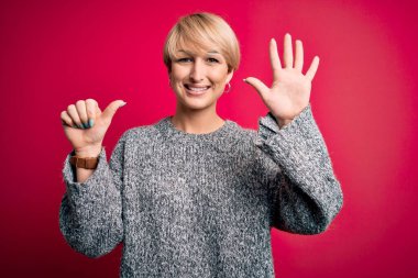 Modern kısa saçlı genç bir kadın pembe arka planda gündelik süveter giyiyor ve altı numaralı parmaklarıyla işaret ederken kendinden emin ve mutlu gülümsüyor..