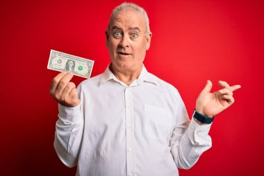 Orta yaşlı, ağarmış bir adam, izole edilmiş kırmızı arka planda bir dolar tutuyor. Elleri ve parmakları kenarda mutlu mesut işaret ediyor.