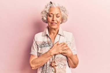 Üstün gri saçlı, günlük kıyafetler giyen, elleri göğsünde, gözleri kapalı gülümseyen ve yüzünde minnet ifadesi olan bir kadın. sağlık kavramı. 